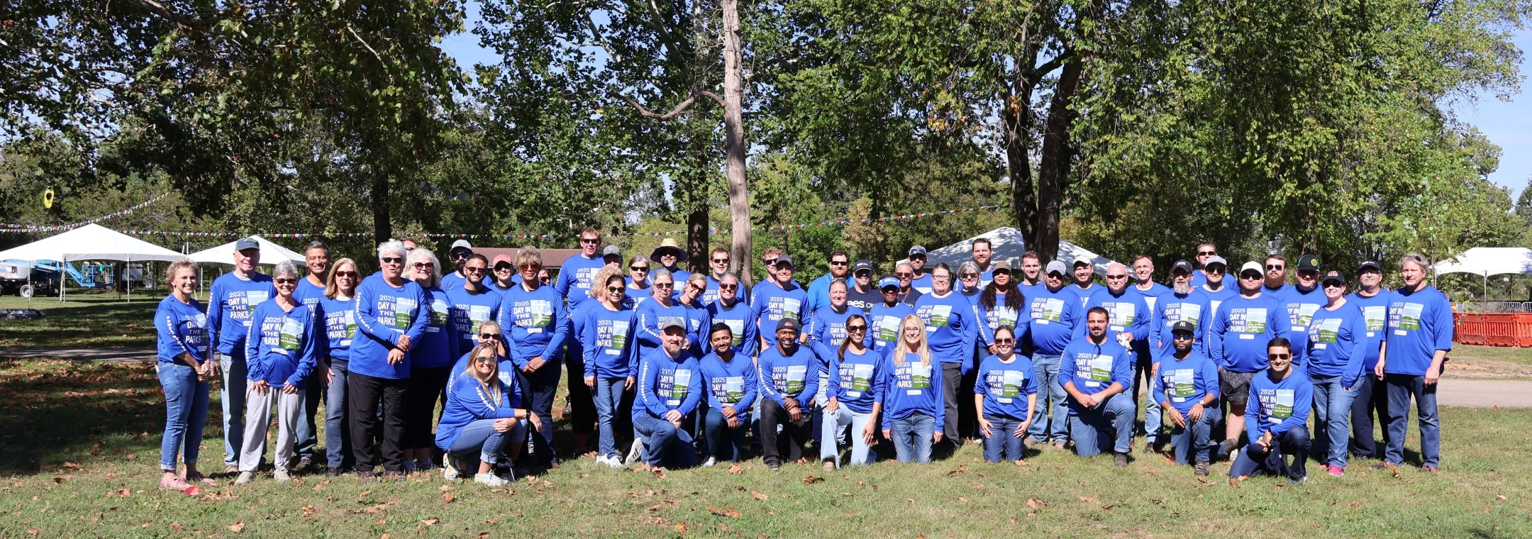 Large group of people wearing matching blue shirts with Day in the Parks printed on them, posing together outdoors in a park on a sunny day, with trees and event tents in the background.