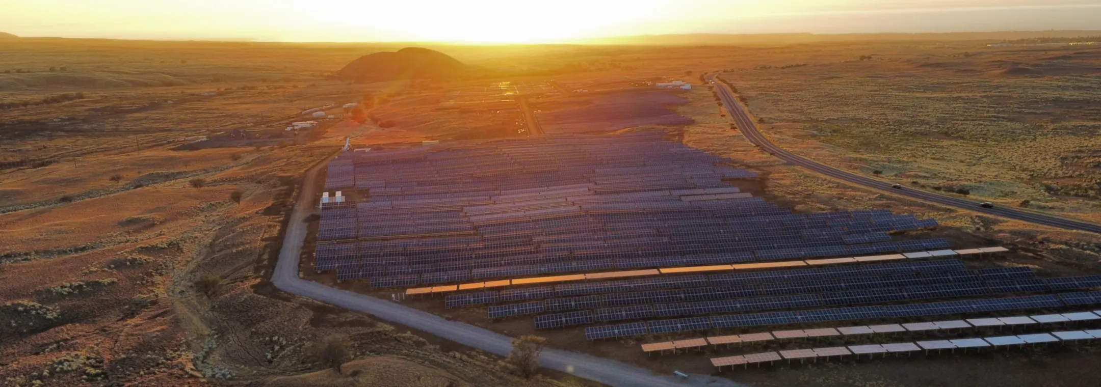 Aerial view of a large solar farm in a vast open landscape at sunset, with rows of solar panels capturing sunlight and a road running alongside the facility.