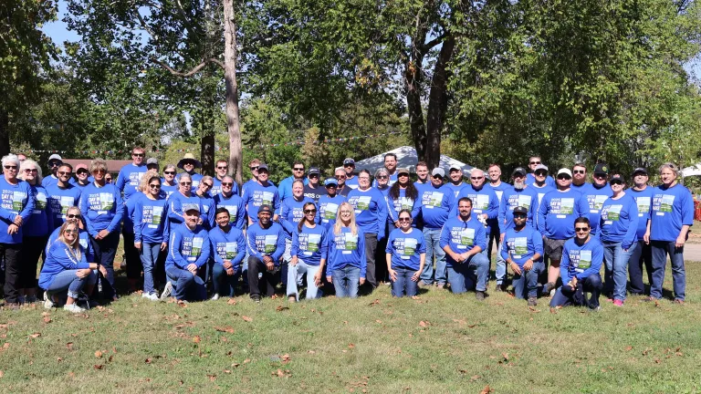 Large group of people wearing matching blue shirts with Day in the Parks printed on them, posing together outdoors in a park on a sunny day, with trees and event tents in the background.