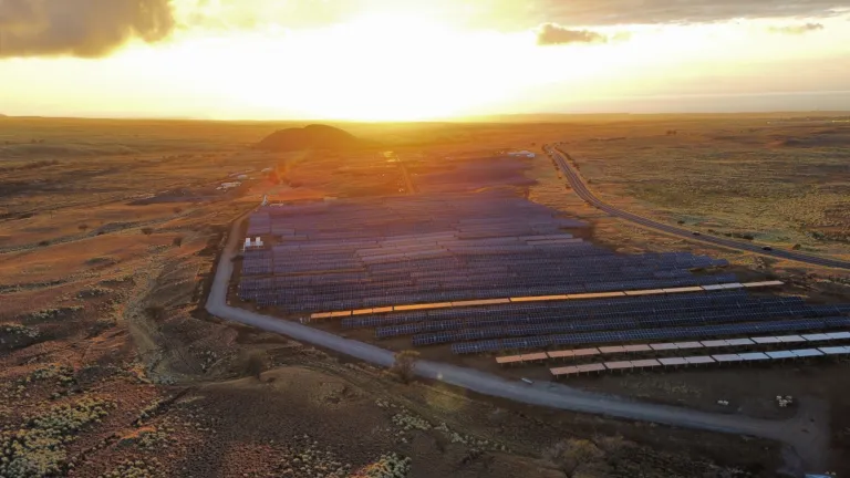 Aerial view of a large solar farm in a vast open landscape at sunset, with rows of solar panels capturing sunlight and a road running alongside the facility.