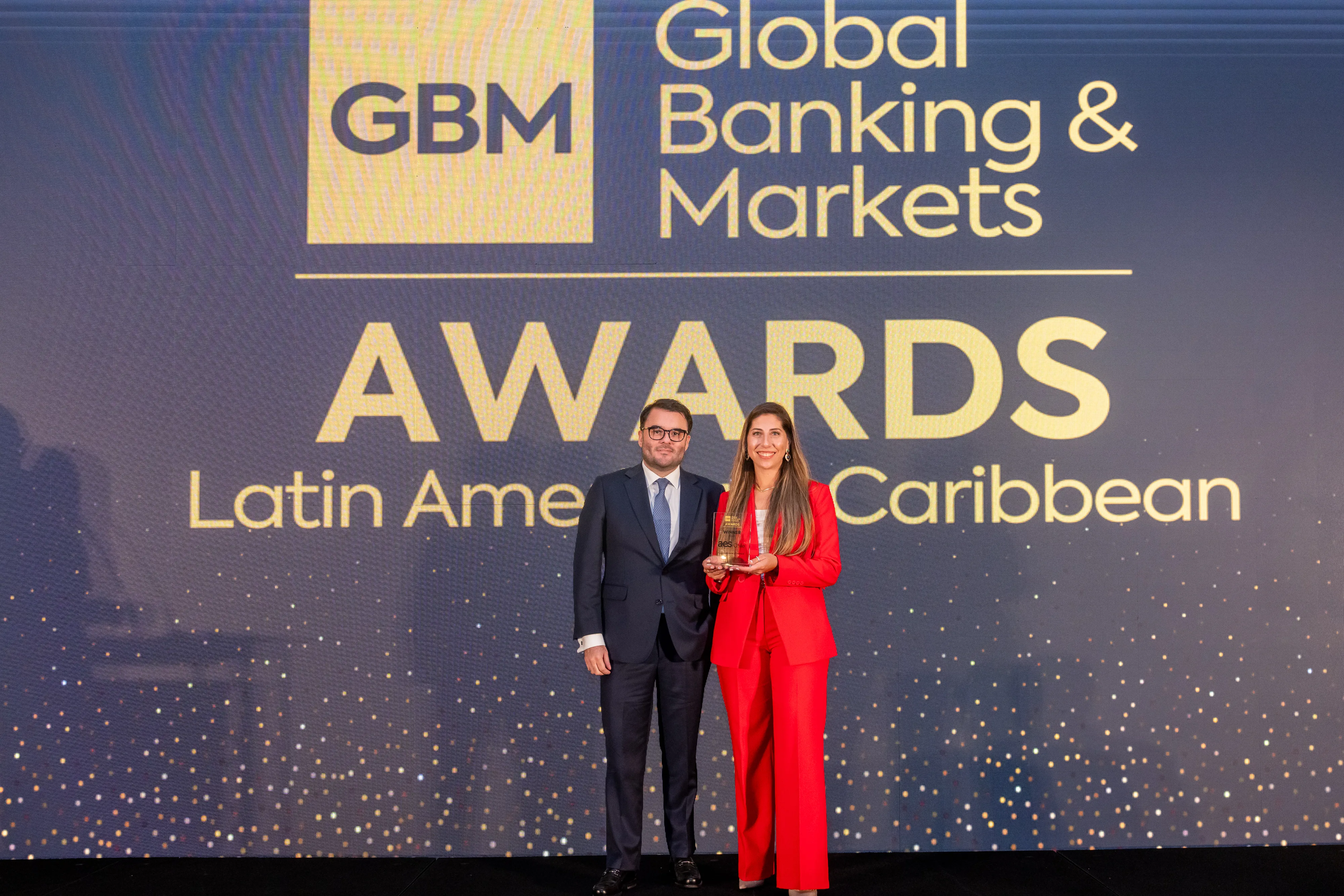 A man in a suit and a woman in a red suit stand on stage holding an award at the Global Banking & Markets Awards for Latin America and the Caribbean, with a large event backdrop behind them.