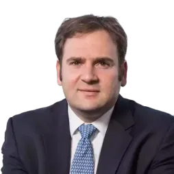 Man in business suit and patterned blue tie sitting in front of a window with a cityscape background, looking at the camera with a neutral expression.